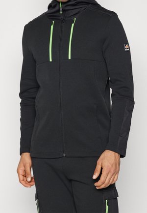 Mand iført sort jakke med lynlås og grønne lynlåsdetaljer samt matchende sorte bukser med grønne lynlåslommer.