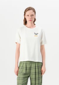 Woman wearing a white t-shirt with a black butterfly and yellow text on chest, paired with green plaid pants, standing against a white background.