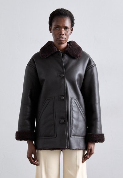 Chaqueta de cuero negra con cuello y puños de piel marrón, con cierre frontal de botones y dos bolsillos laterales. Textura suave y corte holgado.