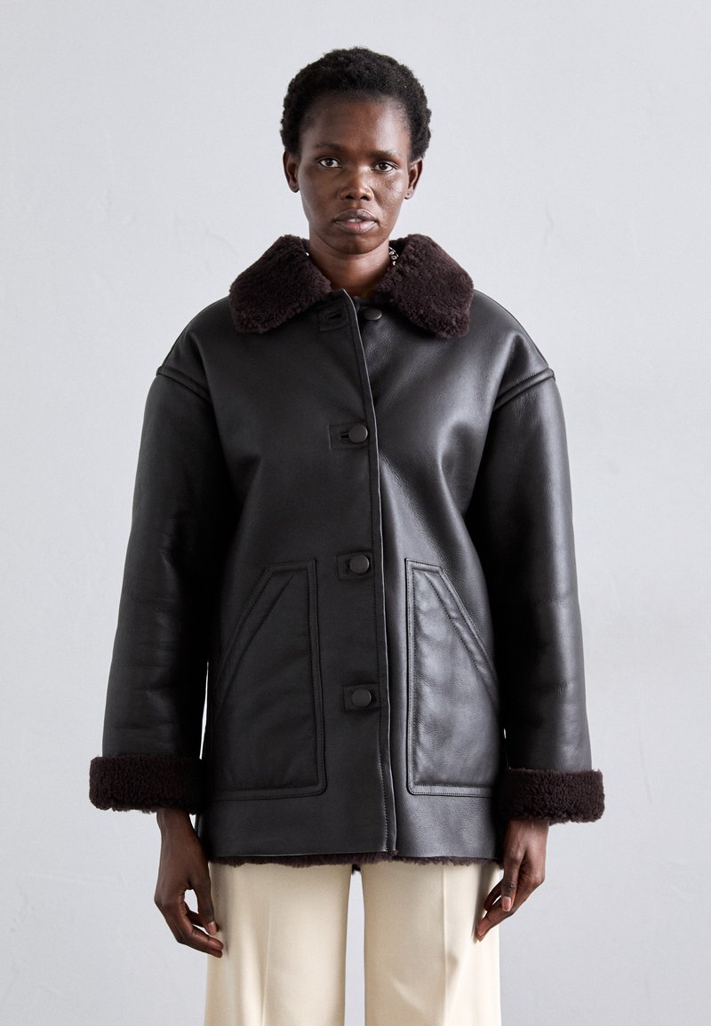 Crna kožna jakna s smeđim ovčjim ovratnikom i manšetama, s prednjim zatvaračem na kopčanje i dva bočna džepa. Glatka tekstura i prostrani kroj.