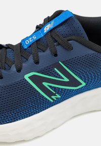 Modrá sportovní obuv s krajkou ze síťoviny, stylizovaným zeleným logem "N", černými šňůrkami a bílou texturovanou podrážkou. Obsahuje modrou visačku s označením "520".