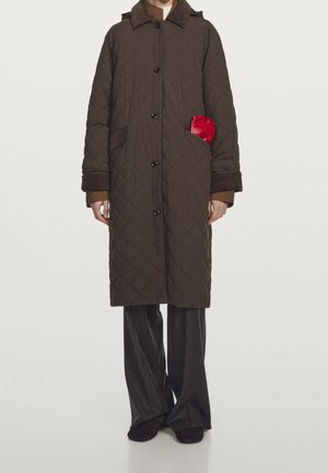 Personne portant un long manteau matelassé marron par-dessus un pantalon large foncé, avec un petit objet rouge visible dans la poche droite du manteau.