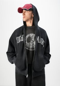 Young person wearing a red cap, black hoodie with "NIGHTMARE" text, and a black t-shirt featuring a skull made of smaller skulls, hands in pockets.