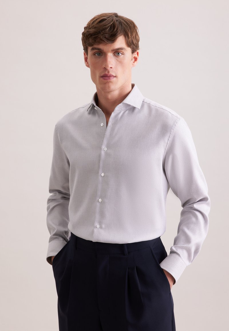 Homme portant une chemise à manches longues boutonnée de couleur gris clair à motif, rentrée dans un pantalon plissé taille haute de couleur bleu marine foncé, les mains dans les poches.