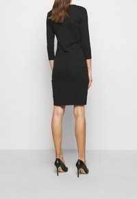 Svart, figurnära klänning med trekvartsärmar och rund halsringning. Bärs med svarta högklackade skor som visar en jämn yta.