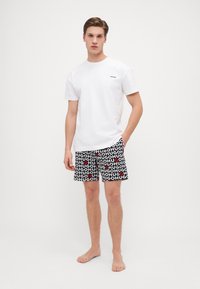 T-shirt en coton blanc avec un petit logo "HUGO" noir, associé à un short noir présentant un motif de texte "HUGO" en blanc et rouge. Le modèle est pieds nus.
