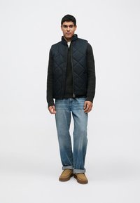 Μπλε καπιτονέ γιλέκο πάνω από σκούρο πουλόβερ πλεκτό, συνδυασμένο με ανοιχτόχρωμο τζιν και καφέ παπούτσια τύπου slip-on. Απλός σχεδιασμός και υφασμάτινα υλικά με υφή.