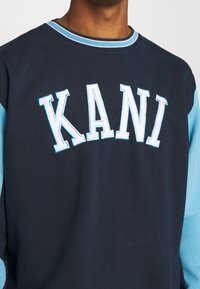 Navy sweatshirt with light blue sleeves, featuring embroidered "KANI" in bold white letters across the chest. Cotton material, crew neck design.
