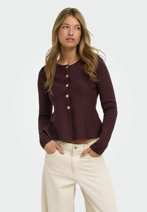 Femme aux longs cheveux blonds portant un pull violet foncé boutonné et un pantalon beige clair, les mains dans les poches, regardant sur le côté.