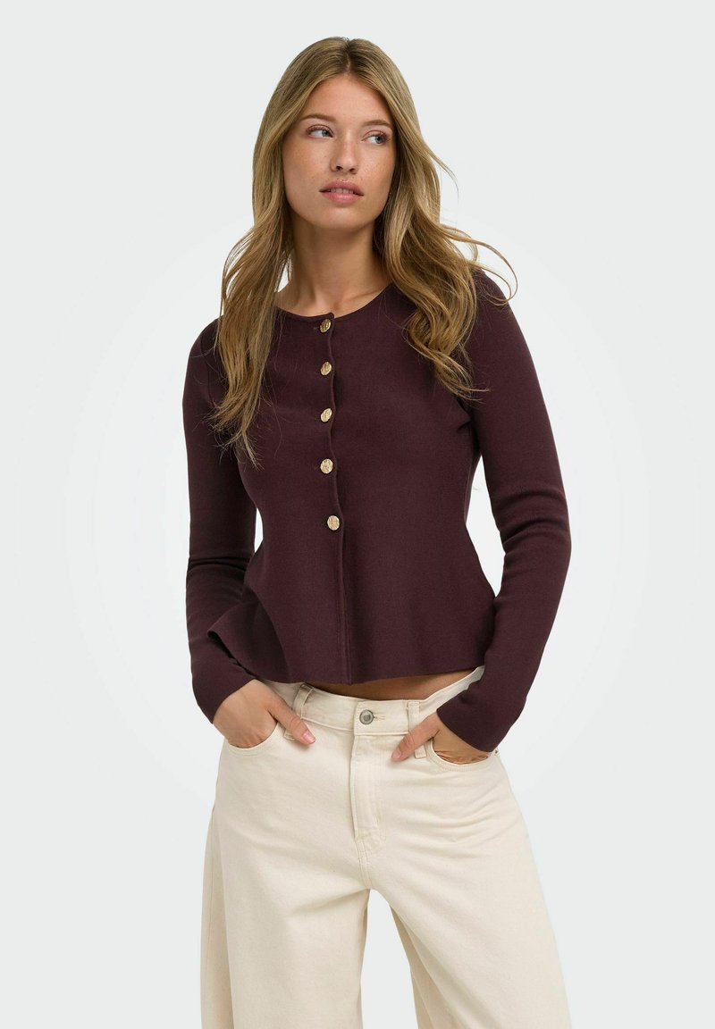 Femme aux longs cheveux blonds portant un pull violet foncé boutonné et un pantalon beige clair, les mains dans les poches, regardant sur le côté.