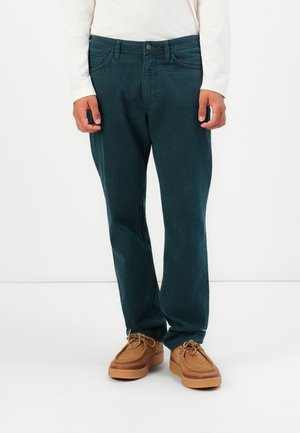 Person in dunkelgrünen, geraden Jeans, weißem Langarmshirt und beigen, geschnürten Mokassins vor weißem Hintergrund.