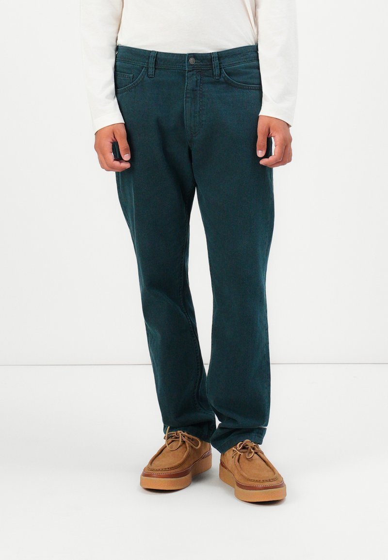 Personne portant un jean droit vert foncé, une chemise blanche à manches longues et des mocassins beige à lacets sur fond blanc.
