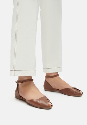 Brązowe skórzane baleriny z odkrytą palcą, z paskiem na kostkę i złożonym detalem z przodu. Noszone z białymi spodniami.