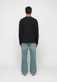 Άνδρας που στέκεται γυρισμένος με την πλάτη, φοράει μαύρο μακρυμάνικο πουκάμισο, ανοιχτό μπλε τζιν παντελόνι με φαρδύ πόδι και γκρι αθλητικά παπούτσια, μπροστά σε ένα απλό φόντο.