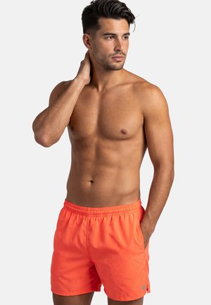 Mężczyzna bez koszulki z krótkimi ciemnymi włosami, ubrany w jasnopomarańczowe spodenki kąpielowe, stojący z jedną ręką na szyi, a drugą w kieszeni.