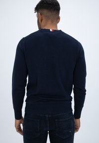 Granatowy sweter z długimi rękawami, ściągaczem na dole i mankietach, z małym czerwono-białym akcentem na tylnej części kołnierza.