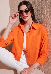 Orange linen button-up shirt with a single pocket, rolled sleeves, worn over a white tank top, paired with white pants and sunglasses.