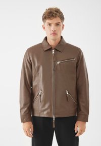 Bruine leren jas met een rits aan de voorkant, puntkraag en twee ritszakken. Heeft een slanke, gladde textuur en een getailleerde pasvorm.