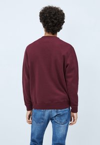 Felpa girocollo color bordeaux con polsini e orlo a coste, realizzata in tessuto morbido. Vista dal retro, abbinata a jeans di denim blu.