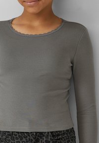 Person, die ein gestreiftes, langärmliges graues Shirt mit einer Spitzenbesatz-Ausschnitt trägt und dunkle Leopardenmuster-Hosen, leicht lächelnd.