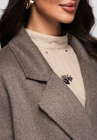 Manteau en laine marron avec col cranté, superposé à un chemisier beige doté d'un col froncé et orné d'une broche florale noire.