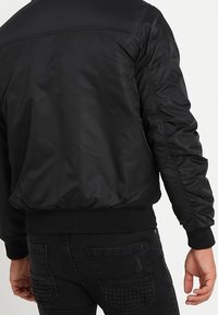 Černá bomber bunda s hladkým, lesklým povrchem, žebrovanými rukávy a lemem, a bočními kapsami. Zadní strana má jednoduchý, minimalistický design.