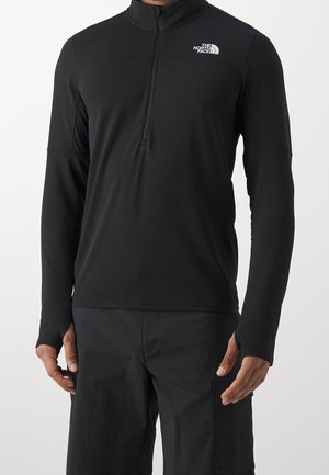 Mies, joka käyttää mustaa pitkähihaista vetoketjullista yläosaa, jossa on The North Face -logo vasemmassa rinnassa, ja mustia housuja sivutaskulla.