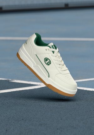 White sneaker with green accents, rubber sole, and perforated toe. Features a logo on the side and textured leather upper.