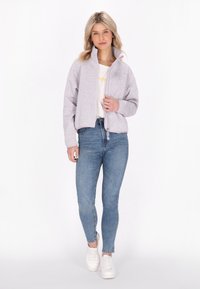 Giacca grigia chiara corta con zip e colletto alto, abbinata a jeans blu aderenti e sneakers bianche. La giacca ha una finitura testurizzata.