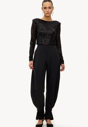 Femme portant un haut à manches longues noir orné de sequins et un pantalon noir taille haute à jambes larges avec des liens à la cheville, debout avec une expression neutre.