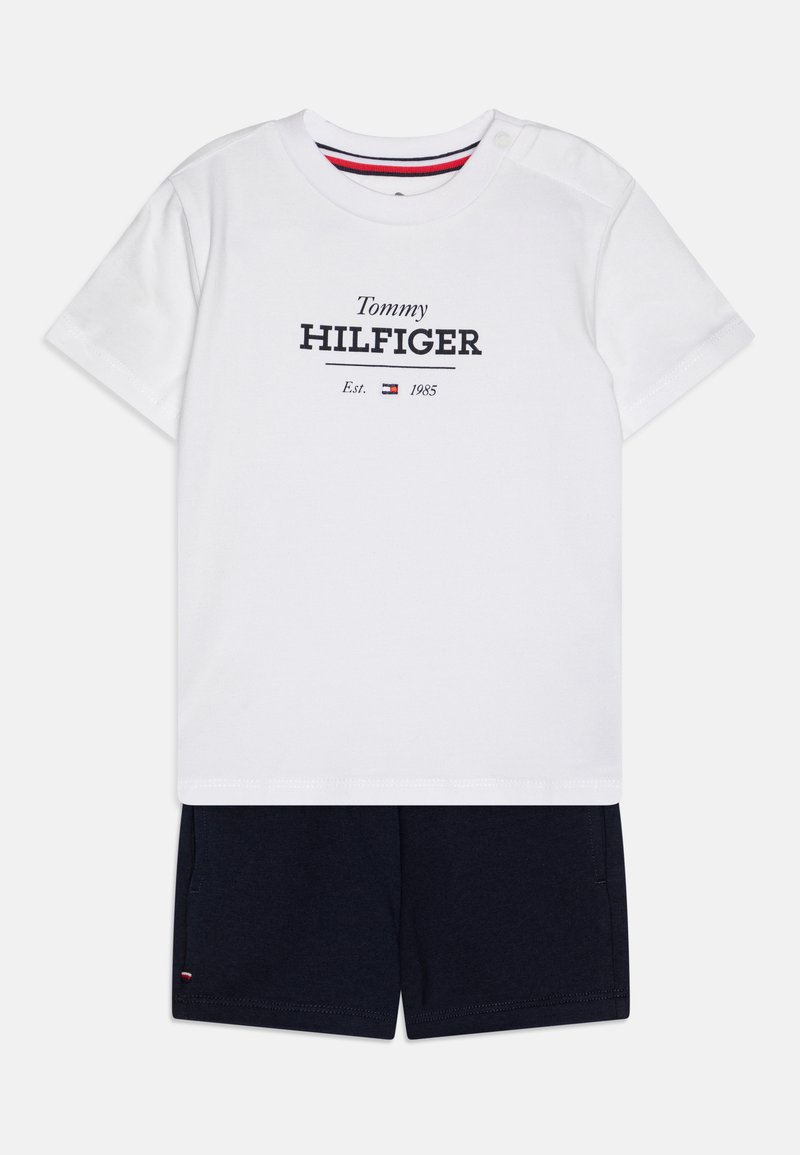 Valkoinen T-paita, jossa on "Tommy HILFIGER" -logo ja tummansiniset shortsit. T-paidassa on lyhyet hihat, pyöreä kaula-aukko ja napitus yksityiskohtana olalla.