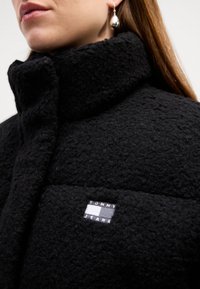 Svart fleece yttertøy med høy krage, med en teksturert finish og et sydd merke som viser "TOMMY JEANS" i hvite og blå striper.