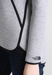 Maglione grigio con bordi neri. Presenta un orlo asimmetrico moderno e il logo "The North Face" sulla manica. Tessuto morbido.
