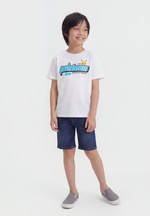 Garçon souriant portant un T-shirt blanc Pokémon avec un dessin de Pikachu, un short en jean et des chaussures grises sans lacets, debout devant un fond uni.