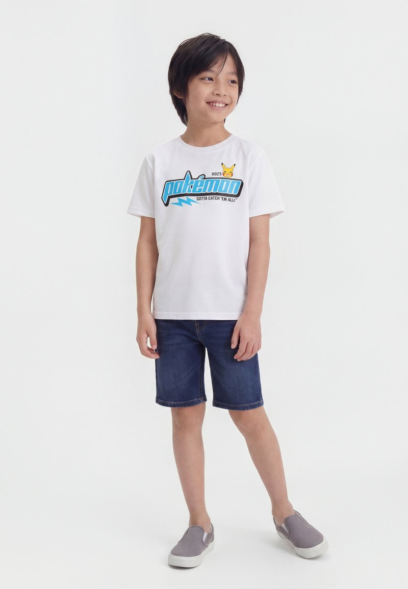 Αγόρι που χαμογελά φορώντας λευκό T-shirt Pokémon με γραφικό του Pikachu, τζιν σορτς και γκρι παπούτσια slip-on, στέκεται μπροστά σε απλό φόντο.
