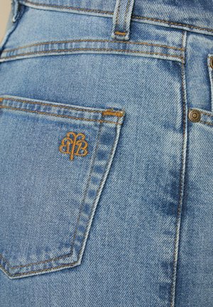 Gros plan sur un jean en denim bleu montrant la poche arrière avec un logo de marque brodé en orange et des détails de couture contrastants.