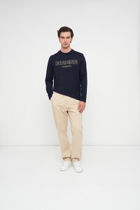 Camicia a maniche lunghe blu navy con logo "GUESS", abbinata a pantaloni beige e sneakers bianche, il tutto su uno sfondo bianco semplice.