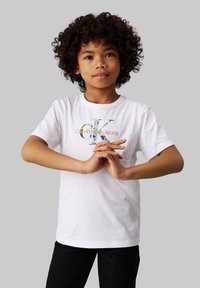 Camiseta de algodón blanca con un logo "CK" multicolor y el texto "Calvin Klein Jeans". Combinada con pantalones negros, con las manos entrelazadas al frente.