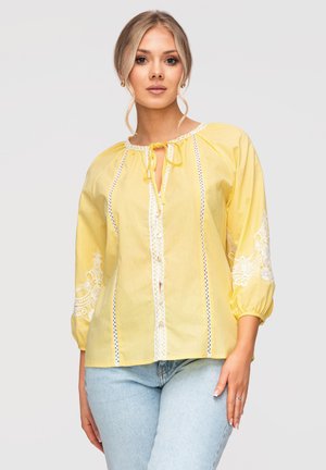 Młoda kobieta w żółtej bluzce z białymi koronkowymi detalami i jasnoniebieskich dżinsach, stojąca na jednolitym tle.