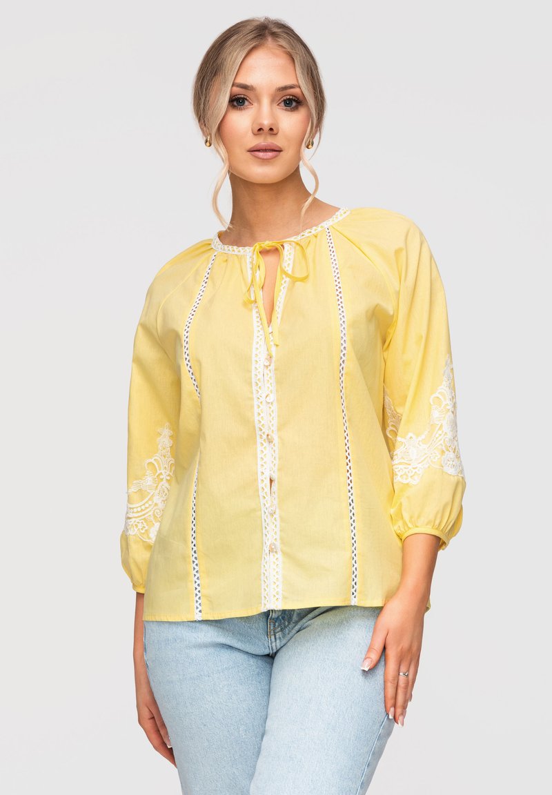 Jeune femme portant un chemisier jaune avec des détails en dentelle blanche et un jean bleu clair, debout devant un fond uni.