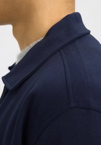 Oldalnézet egy személy nyakáról és válláról, aki sötétkék texturált dzsekit visel világosszürke pulóver fölött.
