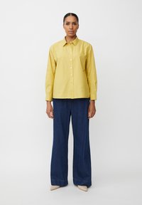 Camicia gialla in cotone con bottoni e maniche lunghe, abbinata a pantaloni in denim scuro dal taglio morbido e scarpe beige a punta.