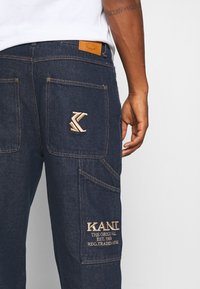 Pantalones de mezclilla azul oscuro con un gran bolsillo trasero que presenta un logo bordado de KANI y el texto "THE ORIGINAL EST. 1989 REG. TRADEMARK" en la pierna derecha.