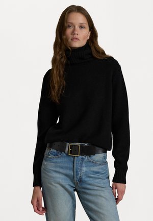 Svart polotröja i mjukt material med ribbat halslinning, tillsammans med ljusblå jeans och ett svart bälte med en guldfärgad spänne.