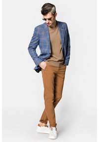 Niebieska marynarka w kratę na brązowym swetrze, dopasowana do brązowych taperowanych spodni i białych sneakersów z beżowymi akcentami. Okulary przeciwsłoneczne dopełniają całość.