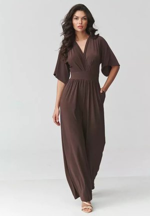 Kobieta o długich ciemnych włosach, ubrana w brązowy kombinezon z szerokimi nogawkami i półrękawami oraz beżowe odkryte buty na obcasie, stoi na jednolitym tle.