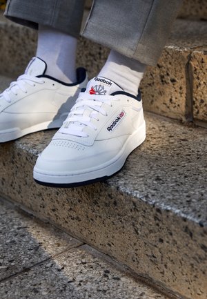 Persoon draagt witte Reebok Classic sneakers met marineblauwe rand en witte sokken, staand op buitenstenen traptreden.