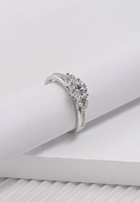 Three-stone silver ring featuring a large central sparkling gemstone, flanked by two smaller stones, with a textured band accentuated by smaller stones.