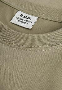Kaki kokvilnas t-krekls ar apaļu kakla izgriezumu un ribotu apkakli, uz kura ir etiķete ar tekstu "R.D.D. ROYAL DENIM DIVISION." Gluda tekstūra.