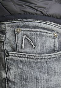 Los jeans de mezclilla gris presentan un bolsillo trasero con un diseño triangular bordado, remaches de metal y detalles de costura en contraste.